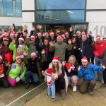 A group of more than 30 people, some in xmas hats, standing in a group and waving at the camera